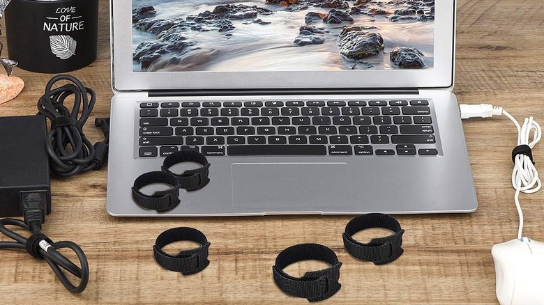 Multiple black Nettbe reusable cable ties, some used on cables and some strewn on a silver laptop and a wooden desk.