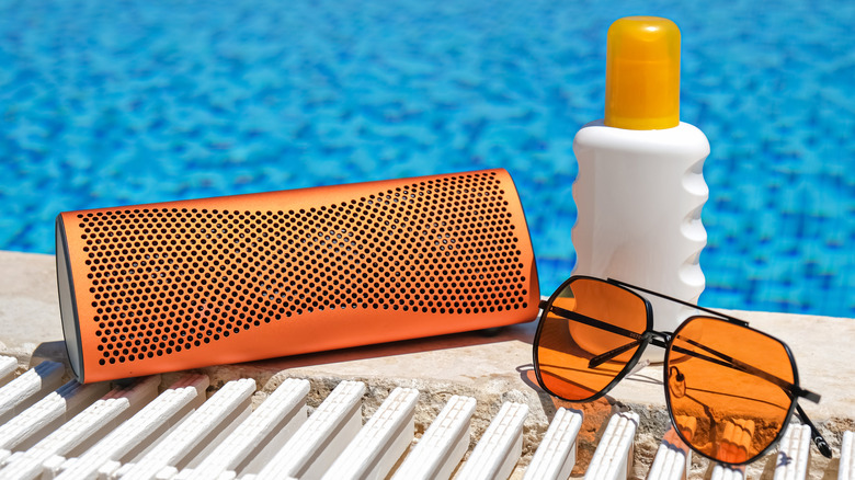 A wireless speaker with sunglasses and a bottle of sunblock beside a swimming pool.