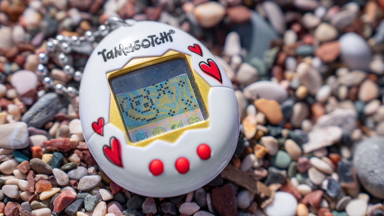 A white Tamagotchi with a red design resting on a pebble beach
