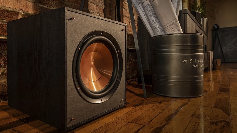 Klipsch R-120SW placed beside a drum containing newspapers on a shiny wooden floor.