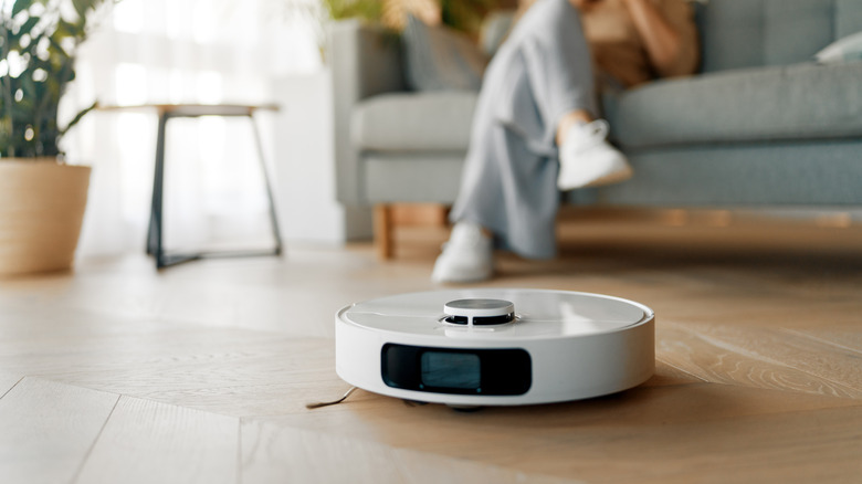 A robot vacuum cleaning the floor while a person is sitting on a couch in the background.