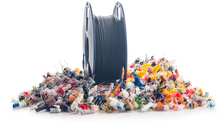 Spool of black 3D printer filament standing upright on a pile of colorful plastic scraps and stringy print waste against a white background.