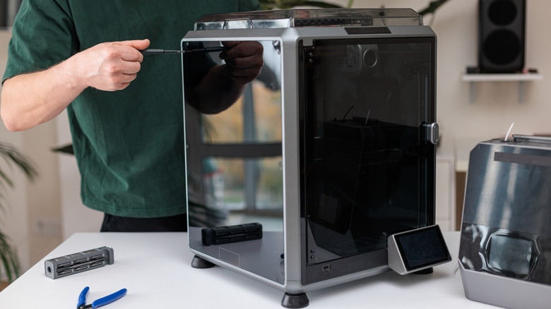 Person using a screwdriver to adjust or assemble an enclosed desktop 3D printer on a work table, with tools and accessories nearby.