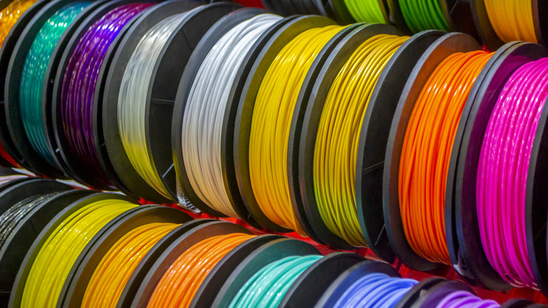 Multiple spools of 3D printer filament in bright colors such as yellow, orange, pink, green, blue, and white arranged side by side on a rack.