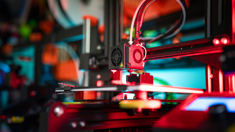 Close-up of a 3D printer extruder head depositing filament onto a print bed, illuminated by red lighting.