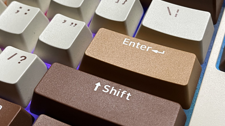 A close-up of custom mechanical keyboard keycaps featuring brown Shift and gold Enter keys with white legends, surrounded by light gray keys and purple backlighting.