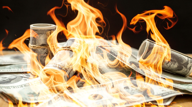 Rolled and scattered U.S. dollar bills engulfed in bright orange flames against a dark background.
