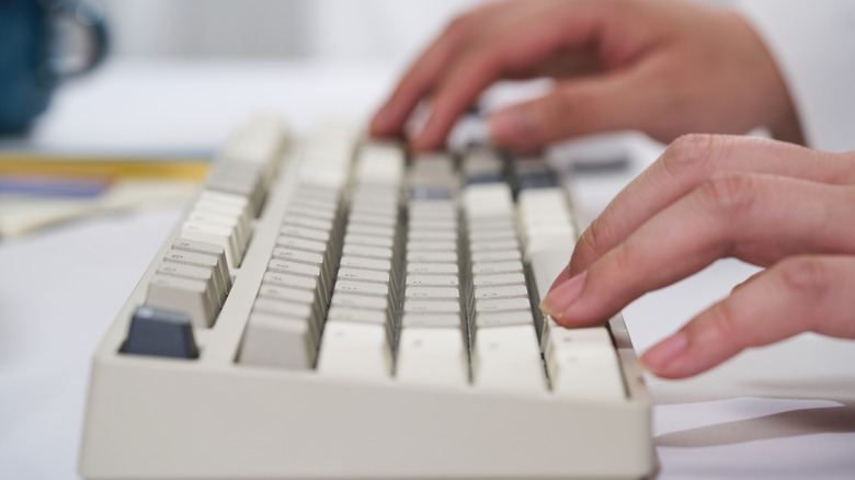 A close-up side view of hands typing on a beige mechanical keyboard on a white desk, with the background blurred.