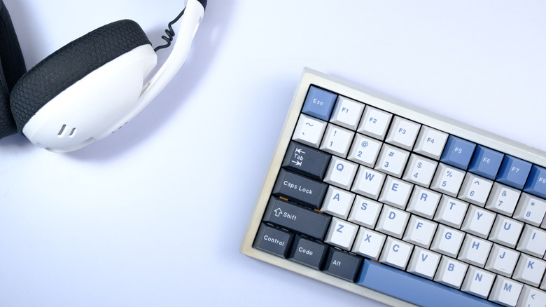 Top-down view of a compact mechanical keyboard with white, blue, and gray keycaps next to a white over-ear gaming headset against a white background.