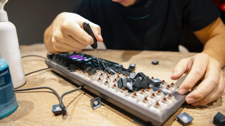 A person using a keycap puller tool to remove keys from a mechanical keyboard during cleaning or maintenance, with loose keycaps scattered across a wooden surface.