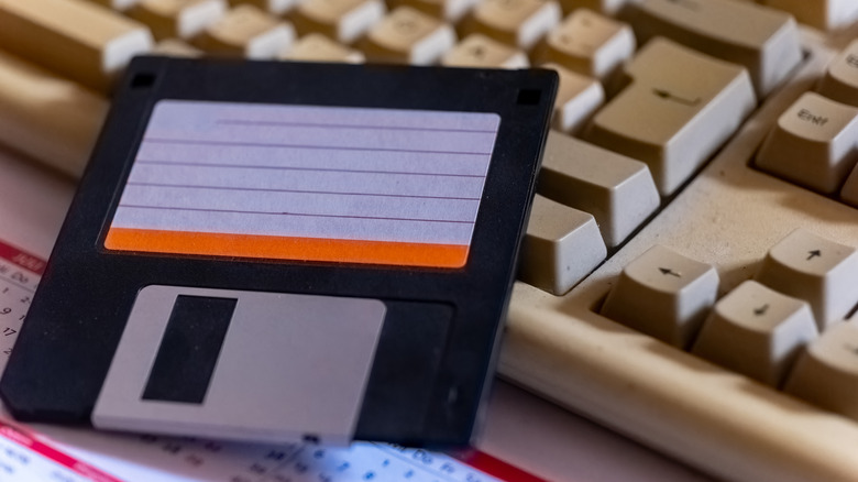 A close-up of a black floppy disk resting on a beige vintage computer keyboard, with retro keys and paper materials visible in the background.