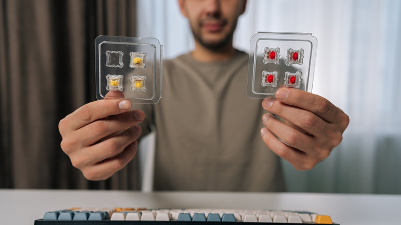A person holding two trays of mechanical keyboard switches (yellow switches in one hand and red switches in the other) above a keyboard on a white surface.