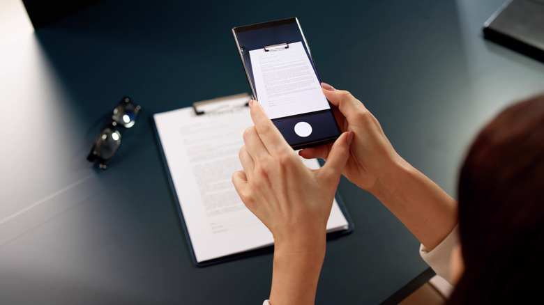 A person using a smartphone to scan or photograph a paper document on a clipboard.