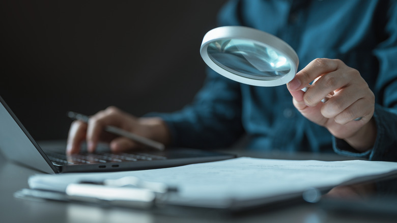 A person holding a magnifying glass while working at a laptop and reviewing printed documents.