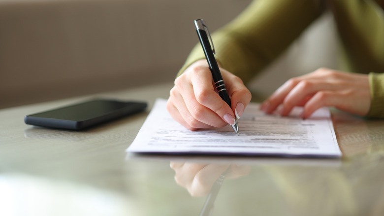 Close-up of a person signing a printed document with a black pen on a desk, with a smartphone resting nearby and the background softly blurred.