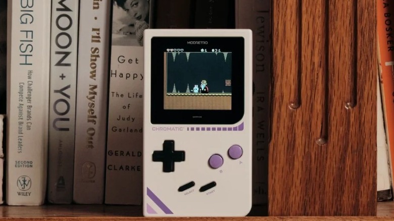 The ModRetro Chromatic, a retro handheld console, sitting on a bookshelf with various books in the background.