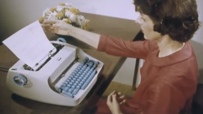 Woman using a vintage typewriter at a desk with a sheet of paper loaded.