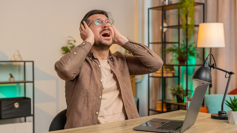A person sitting at a desk with a laptop, covering their ears in apparent frustration.