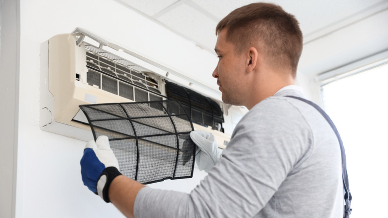 A person wearing work gloves removes a mesh air filter from an open wall-mounted air conditioner.