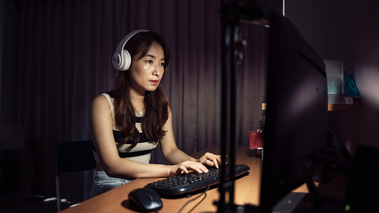 A person wearing over-ear headphones and working at a desktop computer in a dimly lit room.