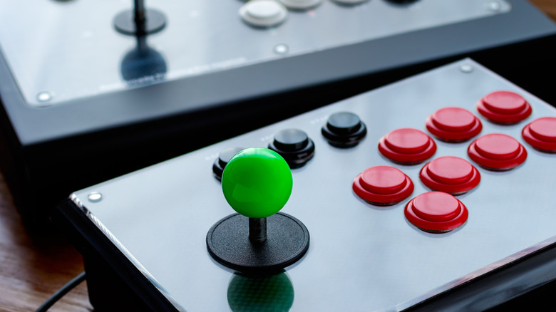 Close-up of a modern arcade fight stick featuring a green joystick and red action buttons, with a second controller blurred in the background.