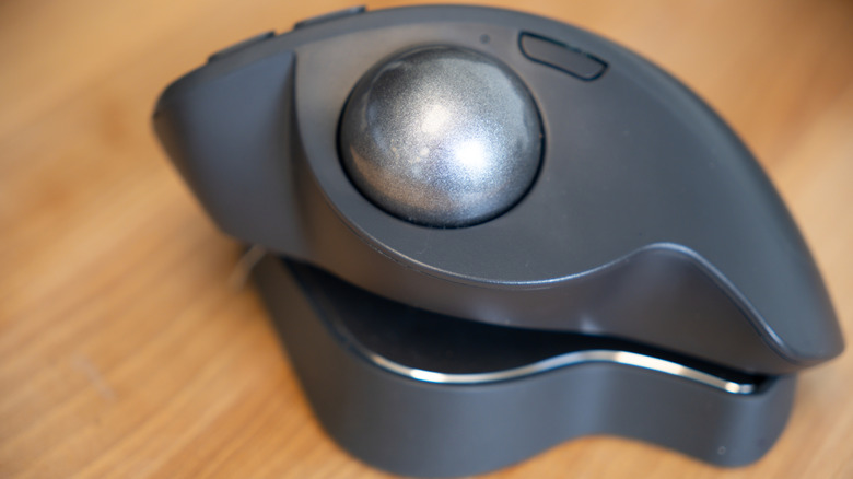 A close-up of an ergonomic trackball mouse with a large metallic trackball and sculpted black body resting on a wooden surace.
