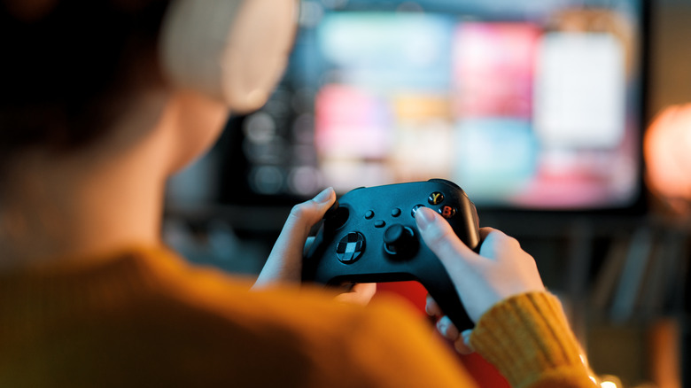A person holds an Xbox controller with a blurred TV visible in the background.