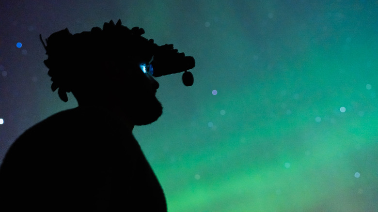 Silhouette of a person wearing night vision goggles against a glowing green and blue night sky.