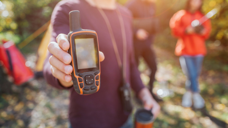 Person holding a satellite phone outdoors at a campsite with blurred campers and tents in the background.