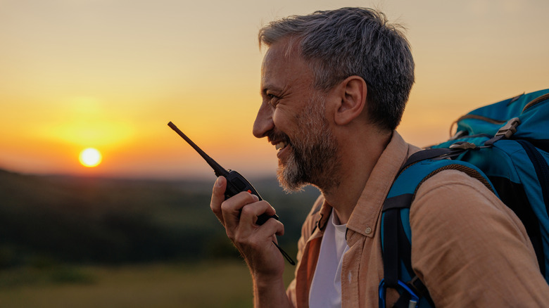 Smiling hiker with a backpack using a handheld walkie-talkie at sunset outdoors.