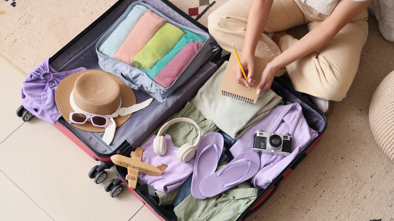 A person writing on a notebook while packing stuff into an open suitcase.