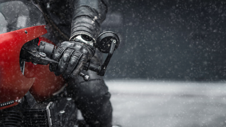 A motorcyclist sits on a bike in snowy conditions with their gloved hand on the clutch lever.