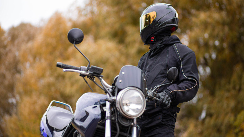 A fully-kitted person stands besides a motorcycle and looks off-camera.