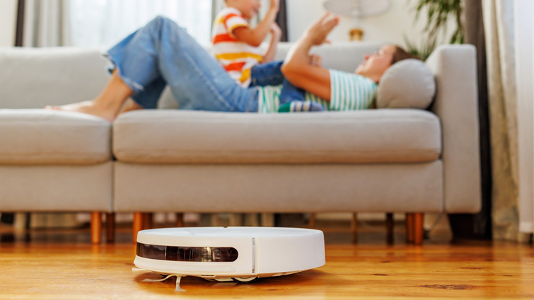 A robot vacuum cleaning the floor while a parent and their child are on the couch in the background.