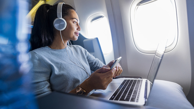 A woman looking out of her window as she's wearing wired headphones and using her phone and laptop.