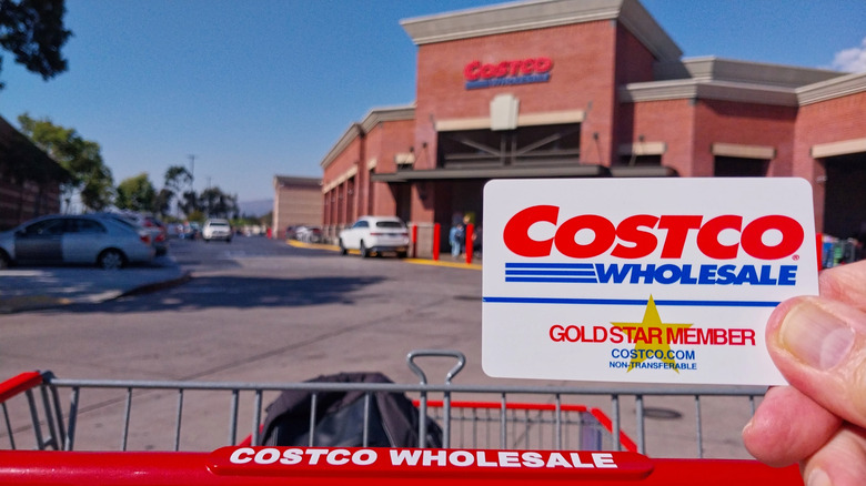 A person holding a Costco membership card with a Costco shopping cart, in front of a Costco store.