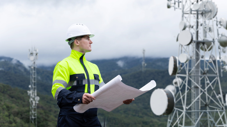 An engineer holds plans while looking at 5G communications towers.