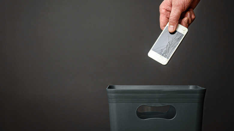 A broken smartphone being dropped into a waste bin