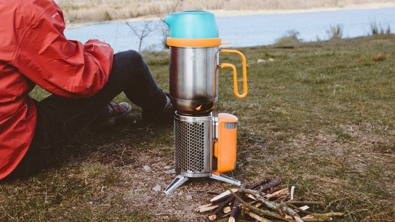 A woman sits next to the BioLite Camp Stove 2 on the ground as it boils water; sticks on the ground will be fuel for the fire
