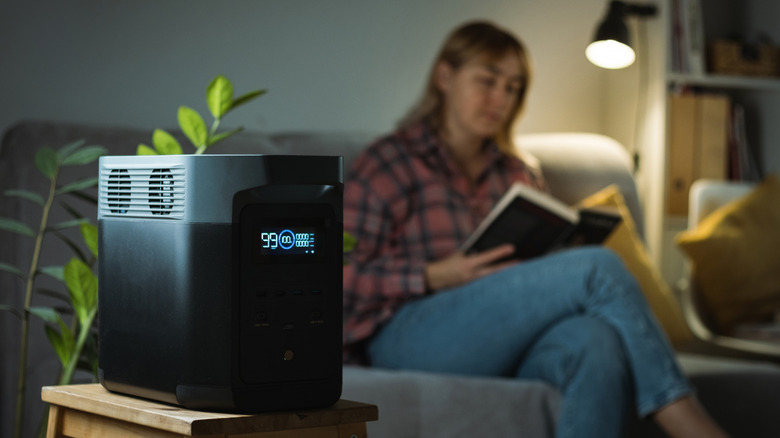 An Ecoflow portable battery in the foreground, with a woman in the background reading a book with a light powered by the battery backup.