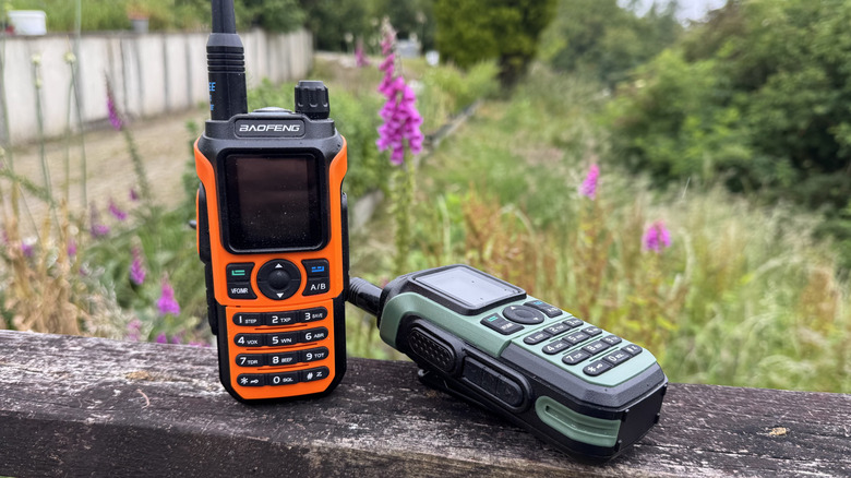 A pair of walkie talkies on a wooden decking in the garden; one green, one orange. Baofeng UV-21R model.
