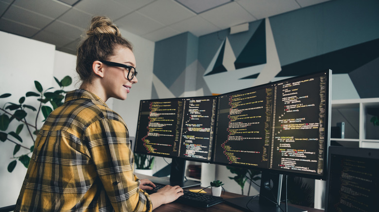 A woman in glasses smiling while looking at code on a dual-monitor setup