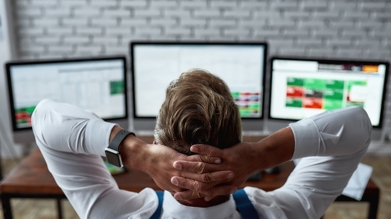 A man relaxing with his hands on the back of his head as he looks at three monitors in front of him