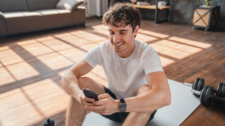 A person sitting on the floor after a workout, looking at their phone.