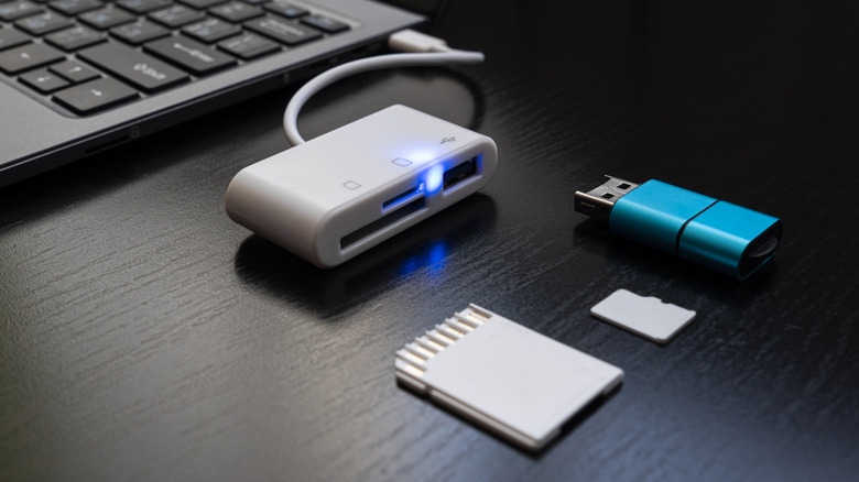 A small white USB dongle plugged into a laptop with an SD card, a microSD card, and a USB drive beside it.