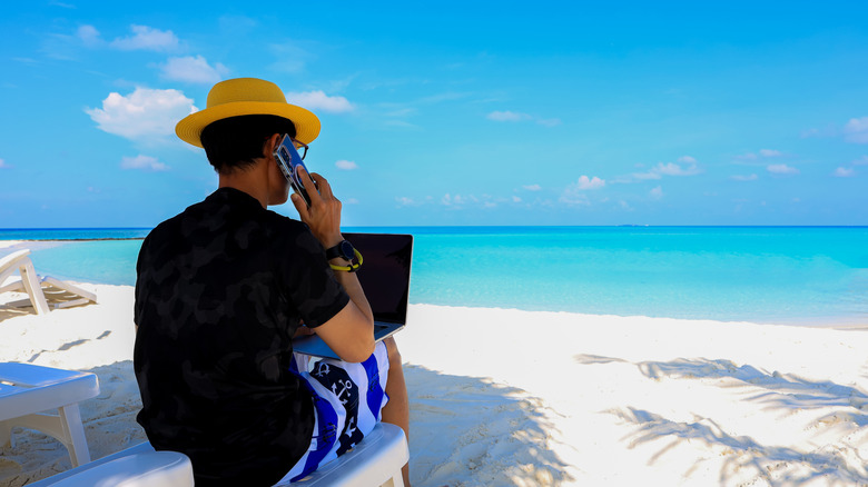 A person talking to someone on the phone while using a laptop and sitting on a lounge chair on a beach.