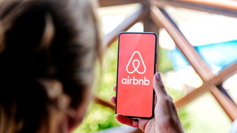 A person holding a phone with the Airbnb logo on the screen with a vacation home in the background.