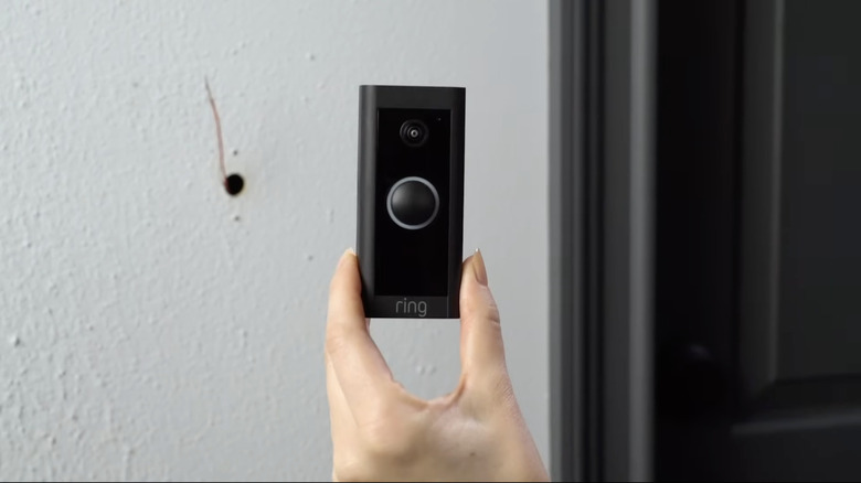 A hand holding black camera doorbell in front of a white wall