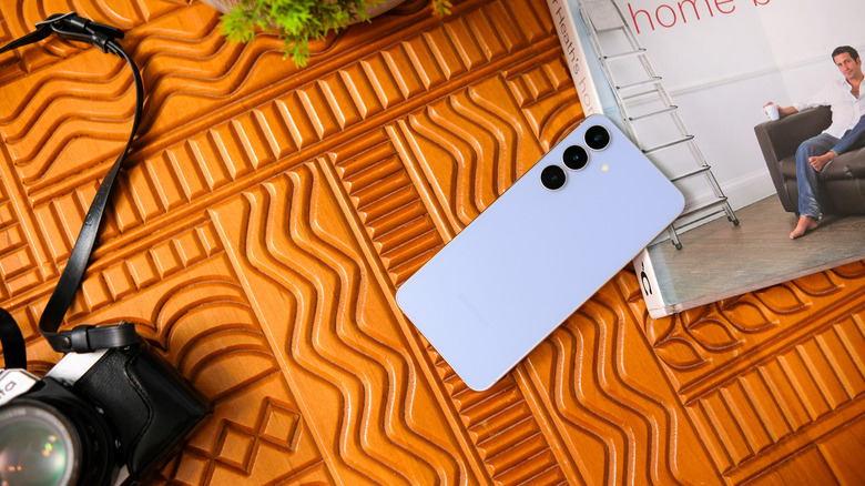 A Samsung Galaxy phone in the center, surrounded by a book, a camera, and a plant partly visible in the backgrond.