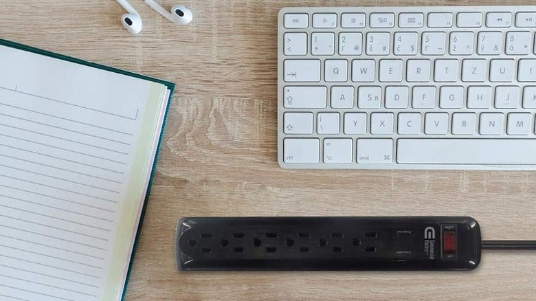 A black Commercial Electric 6-Outlet Surge Protector on a table beside an Apple keyboard, AirPods, and a notebook.